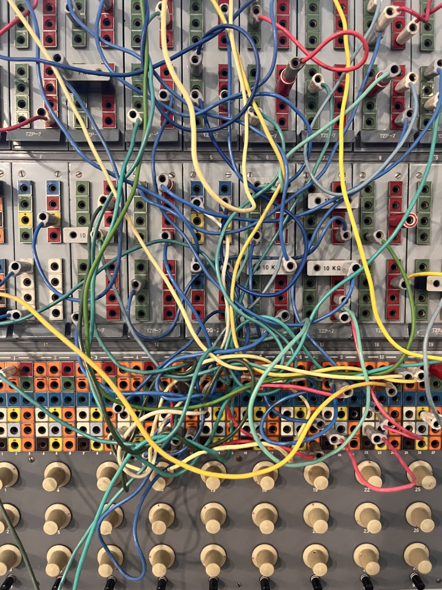 KI generiert: Das Bild zeigt ein komplexes Netzwerk farbiger Kabel, die in verschiedene Steckverbindungen eines analogen Synthesizers oder eines Patchpanels eingesteckt sind. Im unteren Teil sind mehrere Drehknöpfe zu sehen, die wahrscheinlich zur Steuerung des Geräts dienen.
