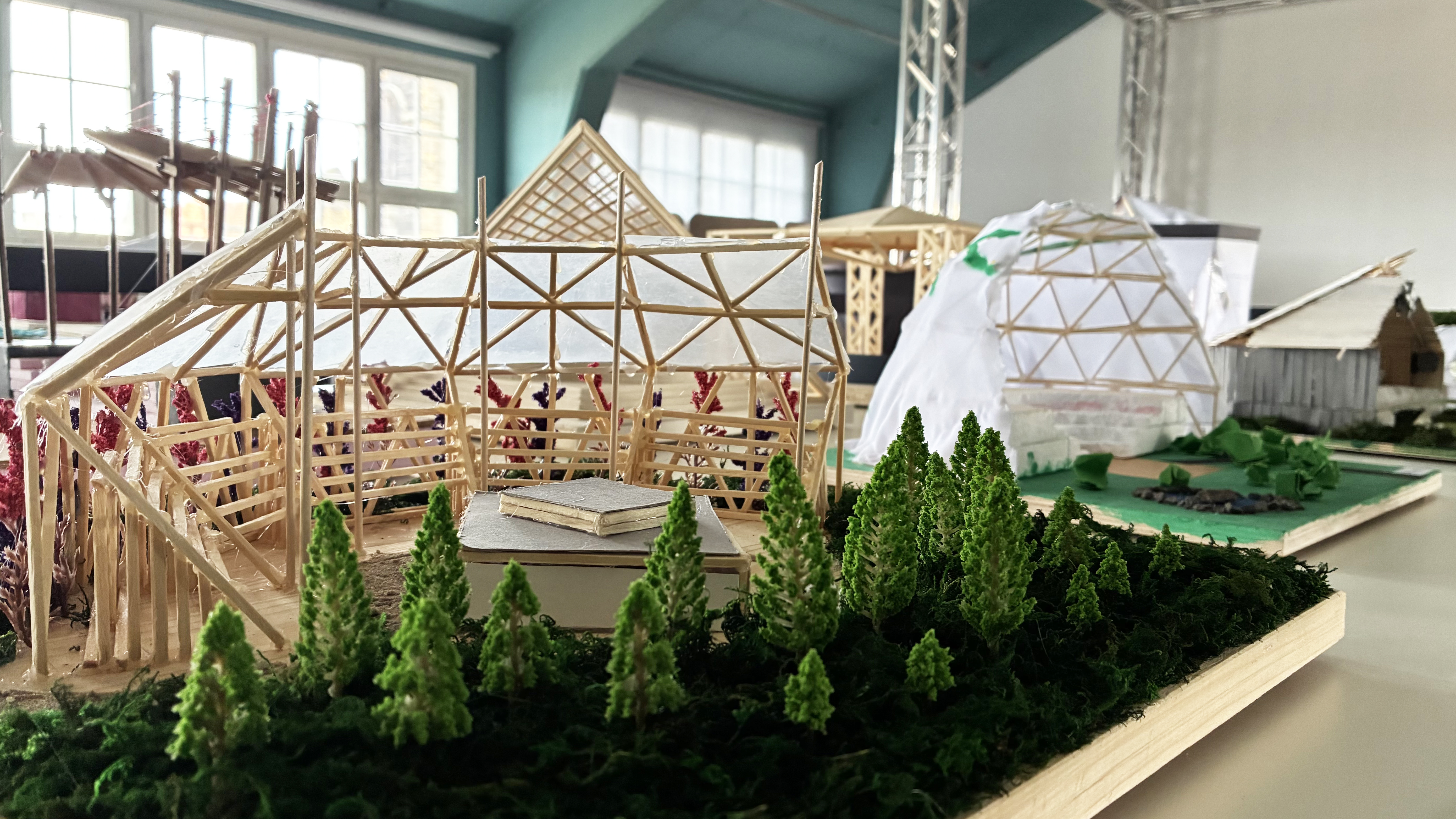 KI generiert: Das Bild zeigt ein Modell einer architektonischen Struktur aus Holz und anderen Materialien, umgeben von Miniaturbäumen und grüner Vegetation. Im Hintergrund sind weitere ähnliche Modellstrukturen zu sehen.