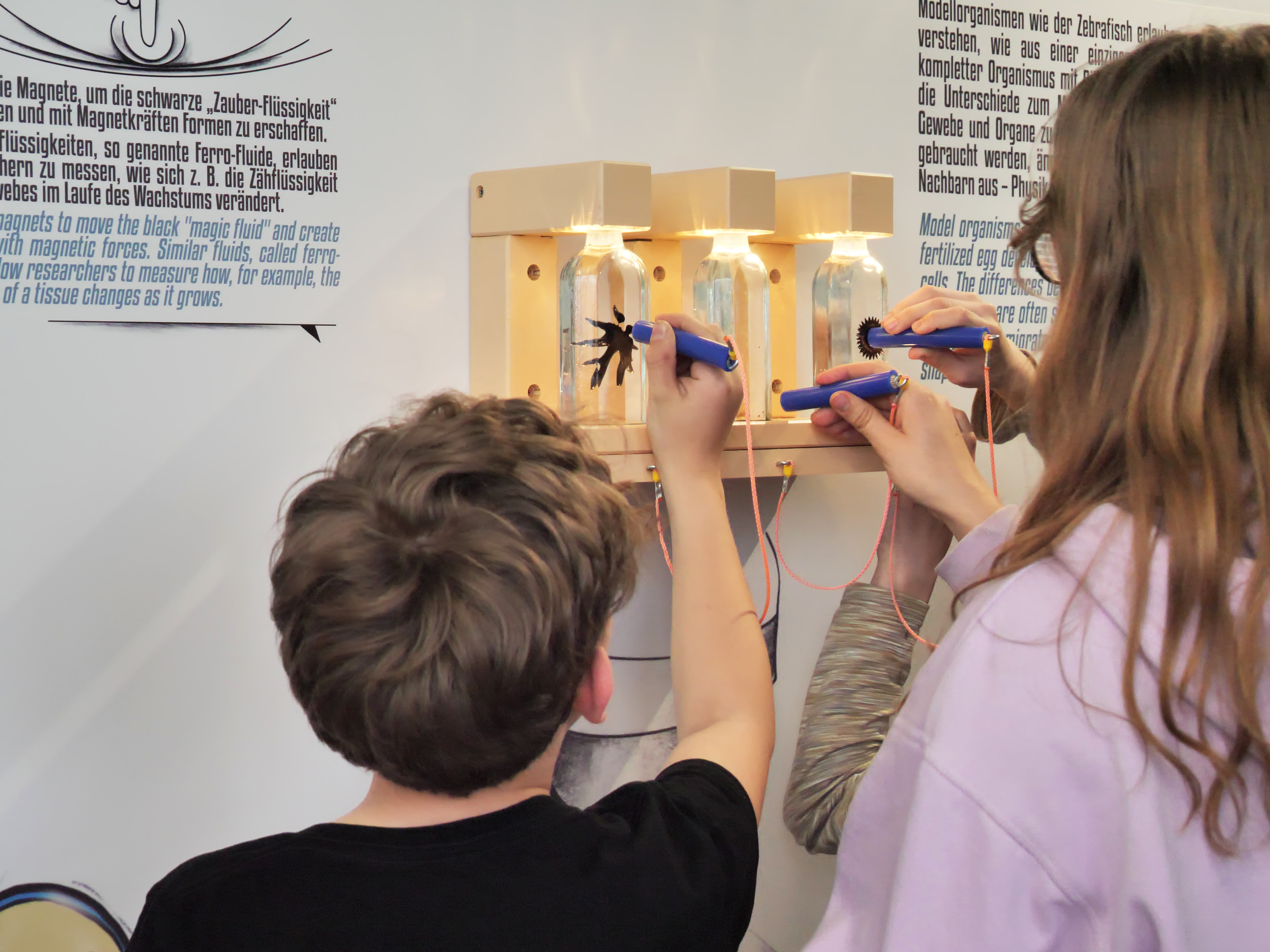 KI generiert: Das Bild zeigt zwei Kinder, die mit Magneten in den Händen vor einer Installation mit Glasflaschen stehen, in denen sich Ferrofluid befindet. Sie experimentieren offensichtlich mit den Magnetfeldern, um die Flüssigkeit zu bewegen und verschiedene Formen zu erzeugen.