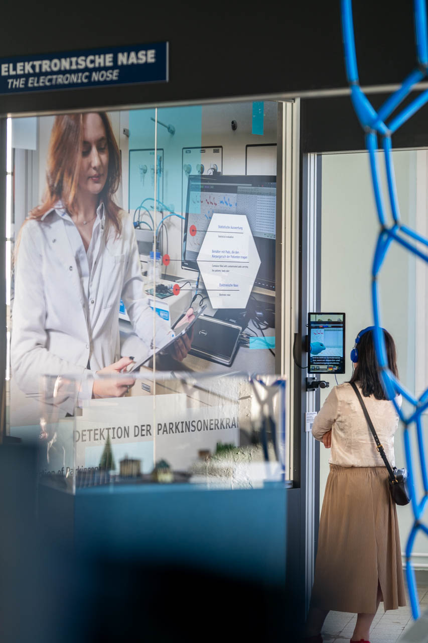 KI generiert: Das Bild zeigt eine Ausstellung oder Präsentation über die "Elektronische Nase", die zur Detektion der Parkinsonkrankheit verwendet wird, mit einem Poster einer Frau in einem Labor. Eine Person, die Kopfhörer trägt, betrachtet die Ausstellung.