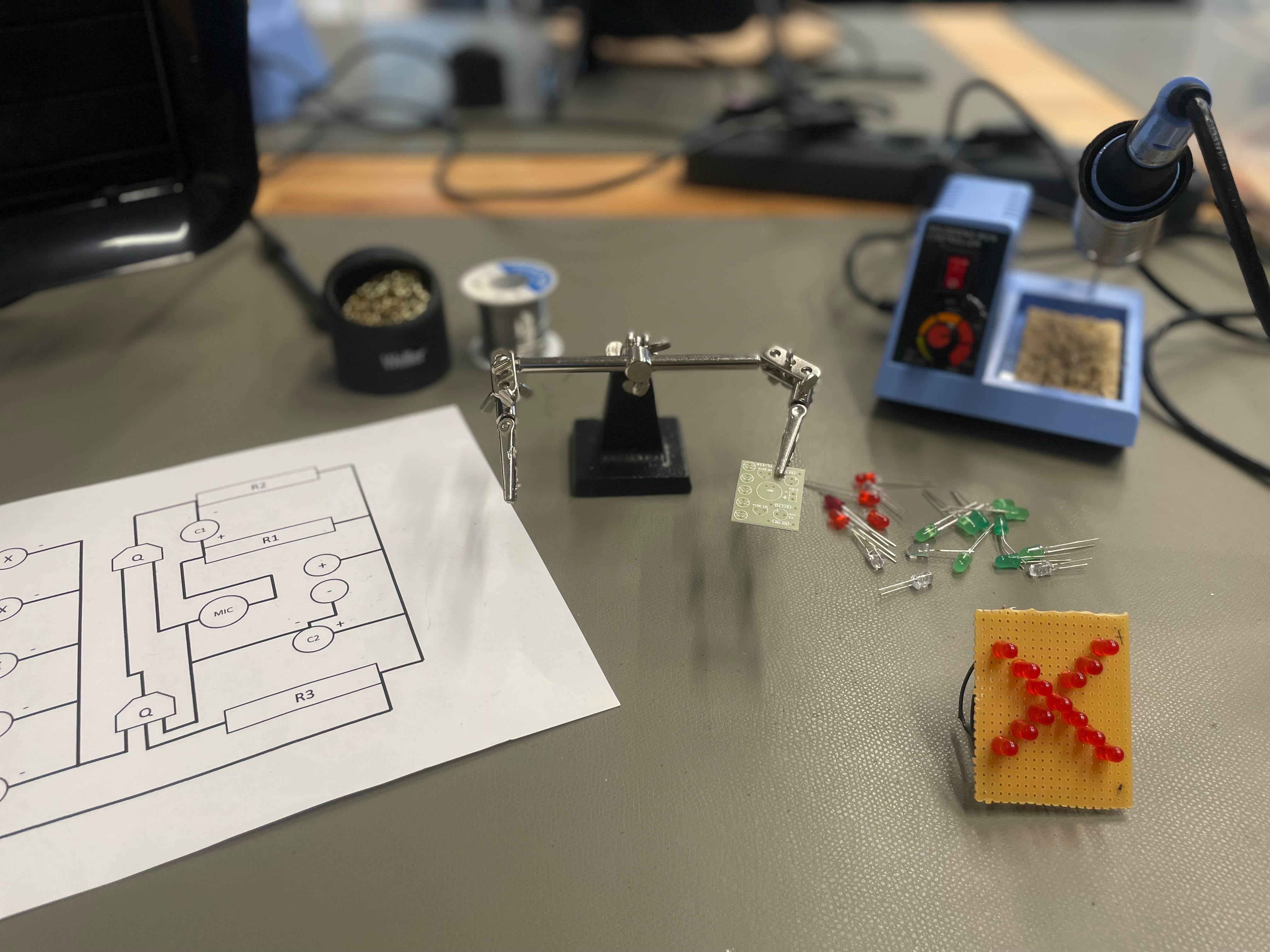 AI generated: The image shows electronic components and tools on a table, including a "third hand" with a circuit board, a circuit diagram as well as soldering utensils and LEDs. The main content appears to focus on the preparation or realisation of a soldering project.