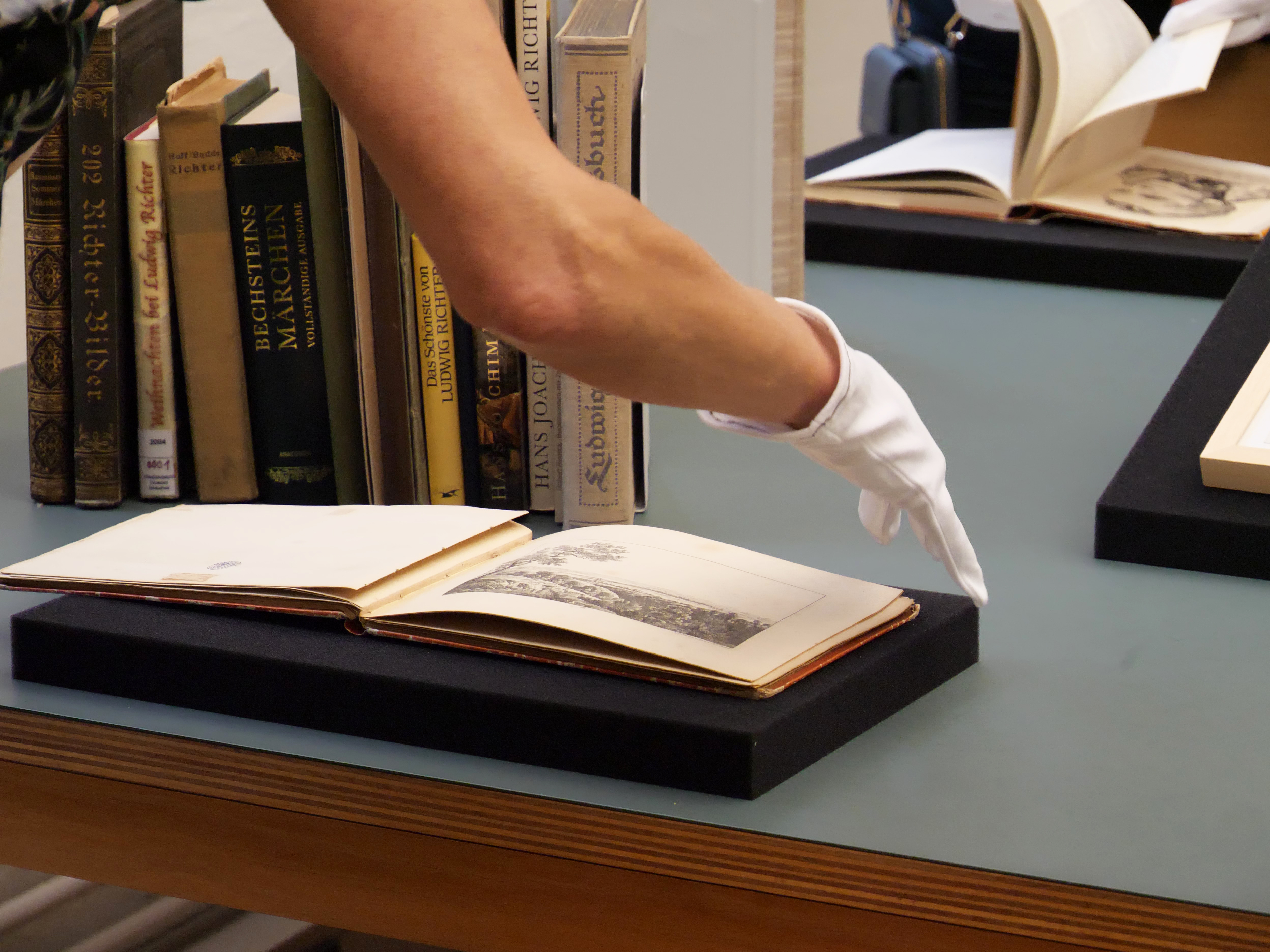 Generated by AI: The image shows a hand wearing a white glove looking at an old, open book lying on a pedestal. In the background, several books are on a shelf.