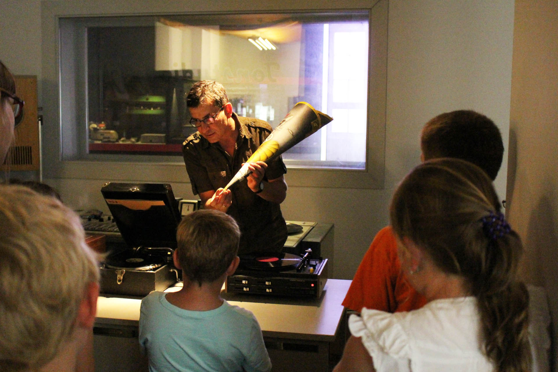 AI generated: A man shows a small group of children a new musical instrument or an old sound player. They all stand around him and seem to be listening curiously.