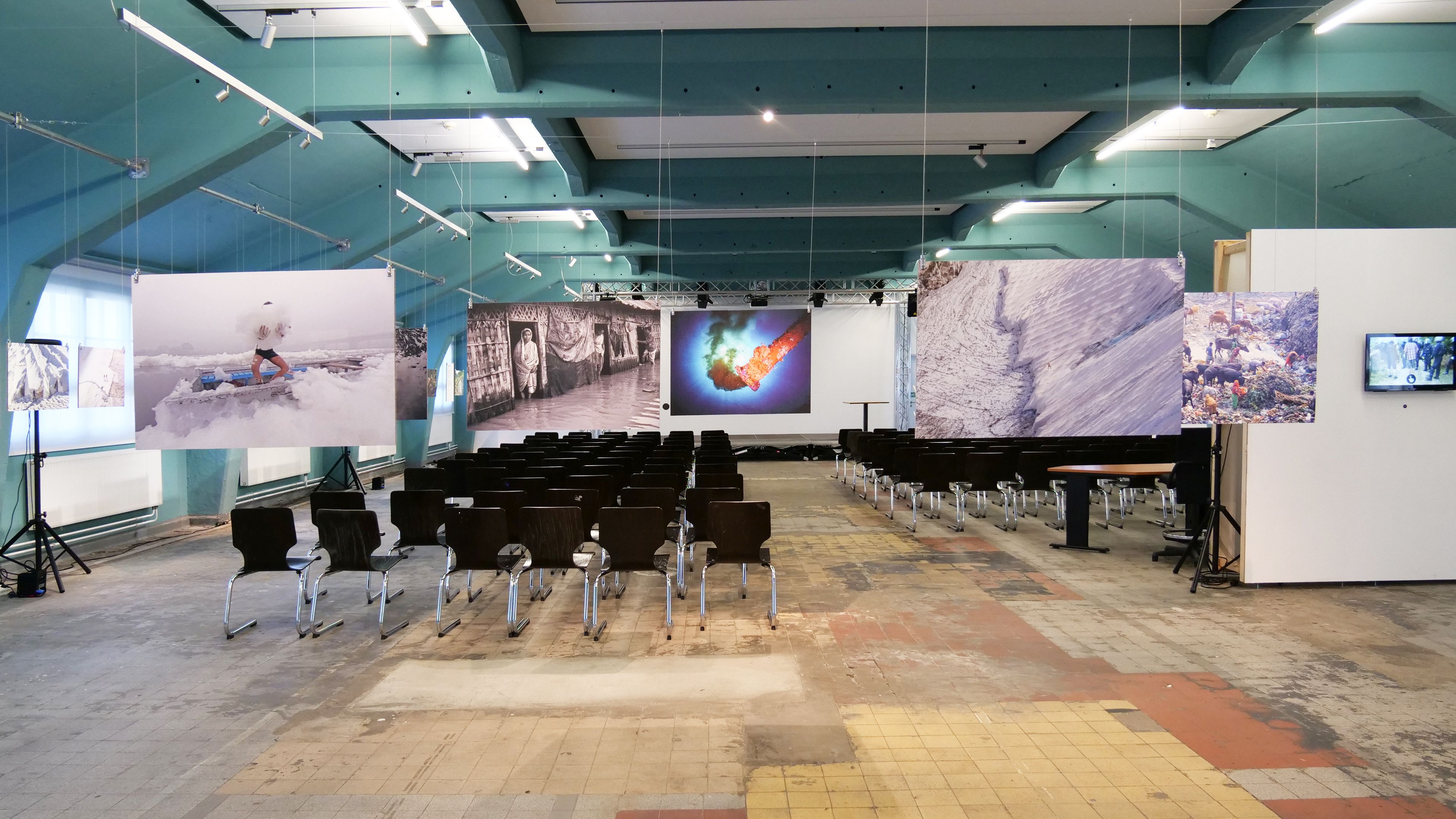 KI generiert: Das Bild zeigt eine Ausstellung mit Fotografien, die in einem großen Raum aufgehängt sind und auf Bildschirmen gezeigt werden. Es gibt mehrere Stuhlreihen vor den Fotografien, die für Besucher bereitgestellt sind.