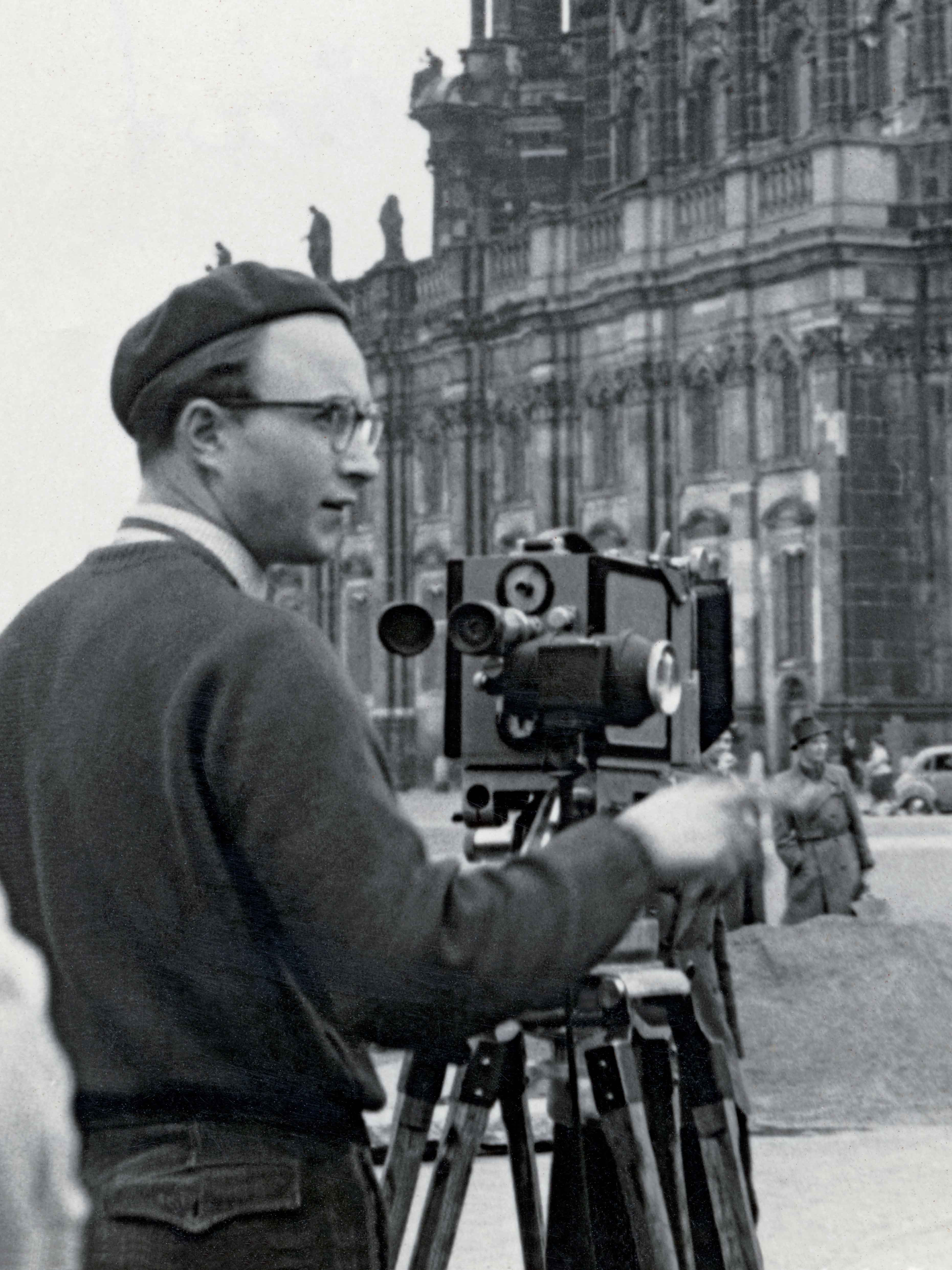 AI generated: The image shows a man standing in front of a large, historic building with a camera on a tripod. The focus is on the filmic or photographic recording of an architecturally impressive structure.
