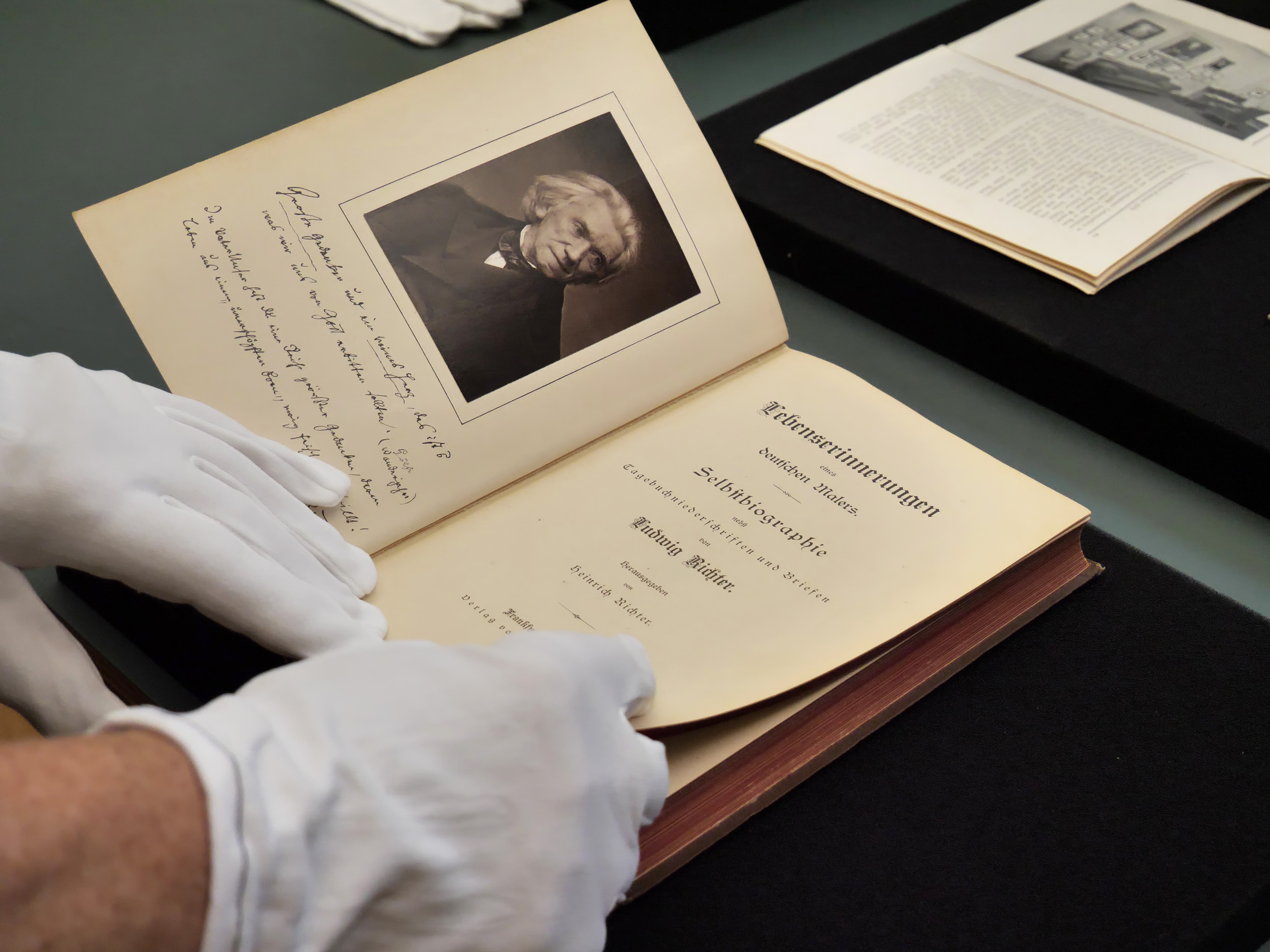 AI-generated: The image shows hands wearing white gloves opening an old book, the pages of which reveal a portrait and a page of text. The main theme is the careful handling of a historical document or book.
