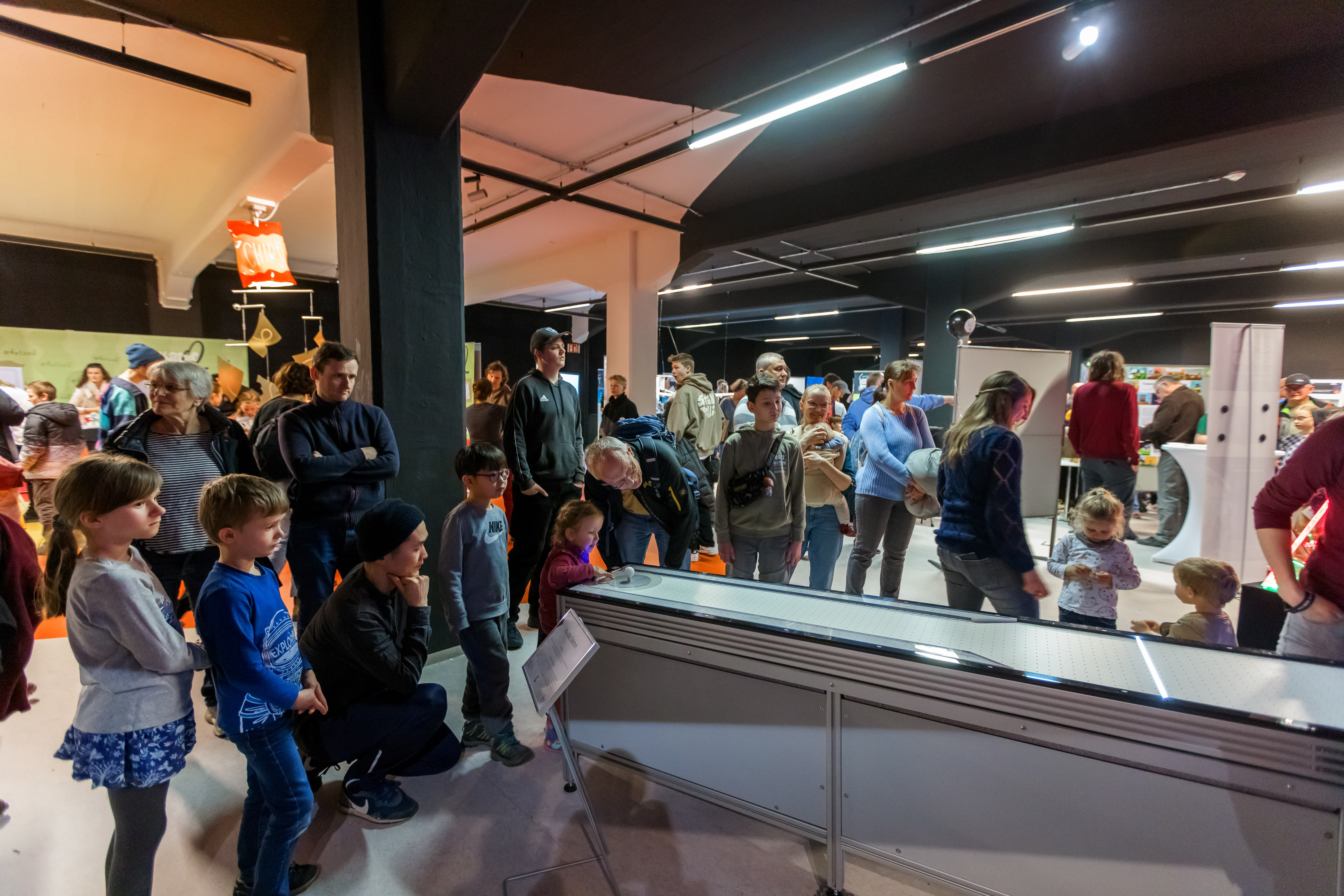 KI generiert: Das Bild zeigt eine Gruppe von Menschen, darunter Kinder und Erwachsene, die sich in einem Raum befinden und eine Ausstellung oder ein interaktives Exponat betrachten. Der Raum wirkt modern und ist mit Beleuchtung ausgestattet, die eine fokussierte und interessierte Atmosphäre schafft.