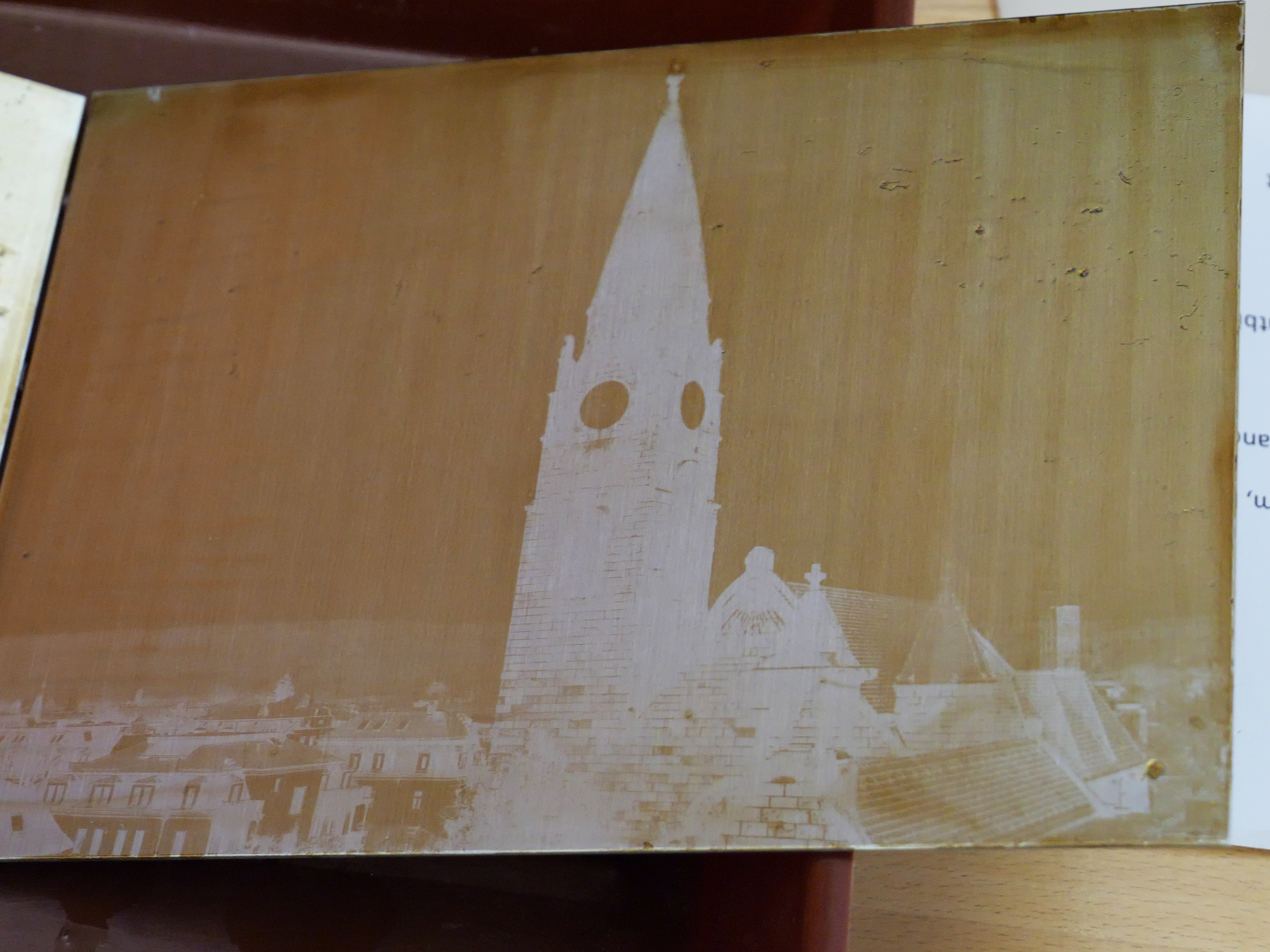 AI generated: The image shows a negative of a photograph of a church tower with a clock and the surrounding cityscape. The image has a sepia-coloured tone and shows architectural details.