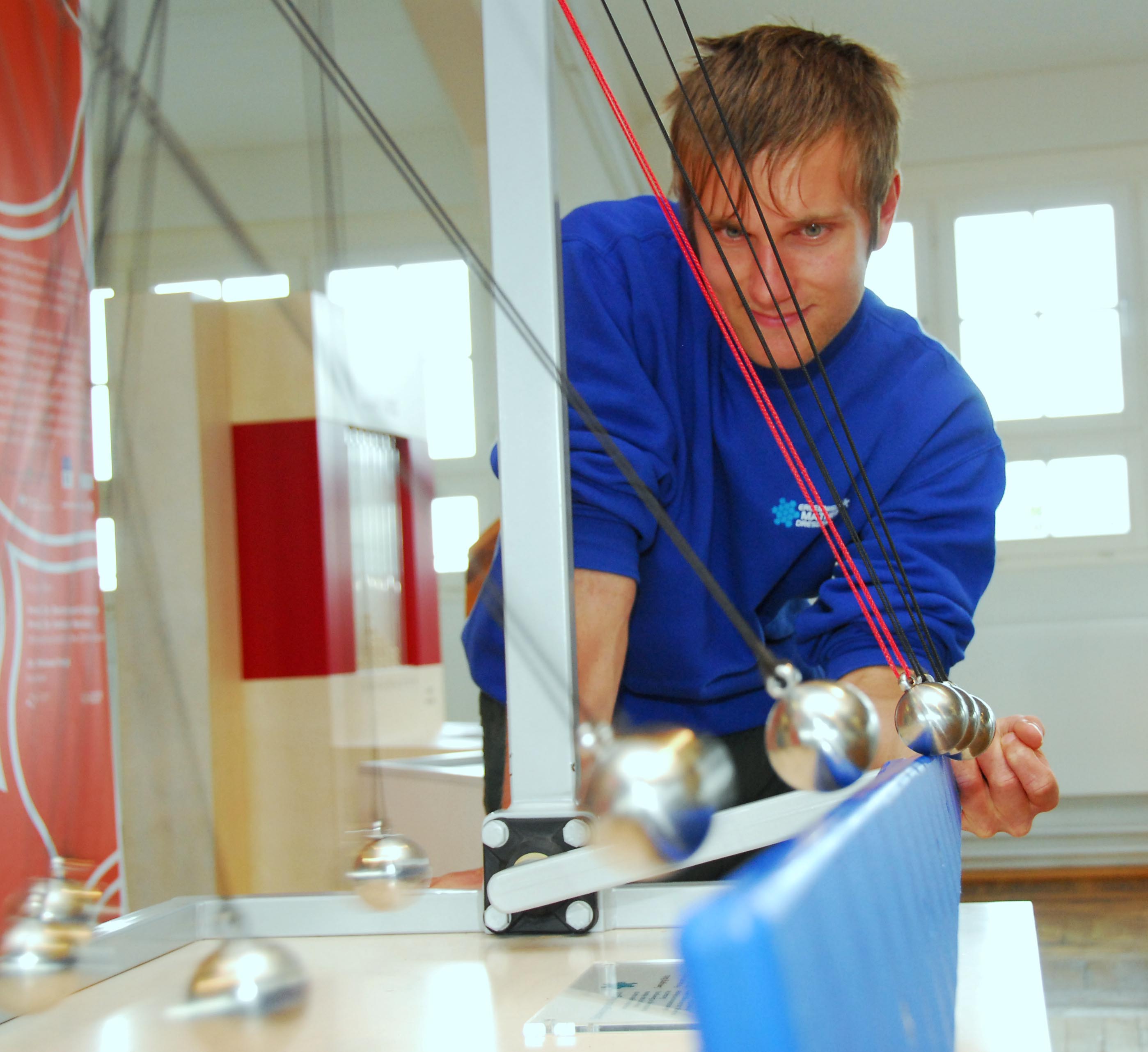 KI generiert: Ein Mann in blauer Kleidung interagiert mit einem Newtonpendel, einem wissenschaftlichen Demonstrationsgerät für Impuls und Energieerhaltung. Die Pendelkugeln schwingen synchron und erzeugen ein faszinierendes visuelles Muster.