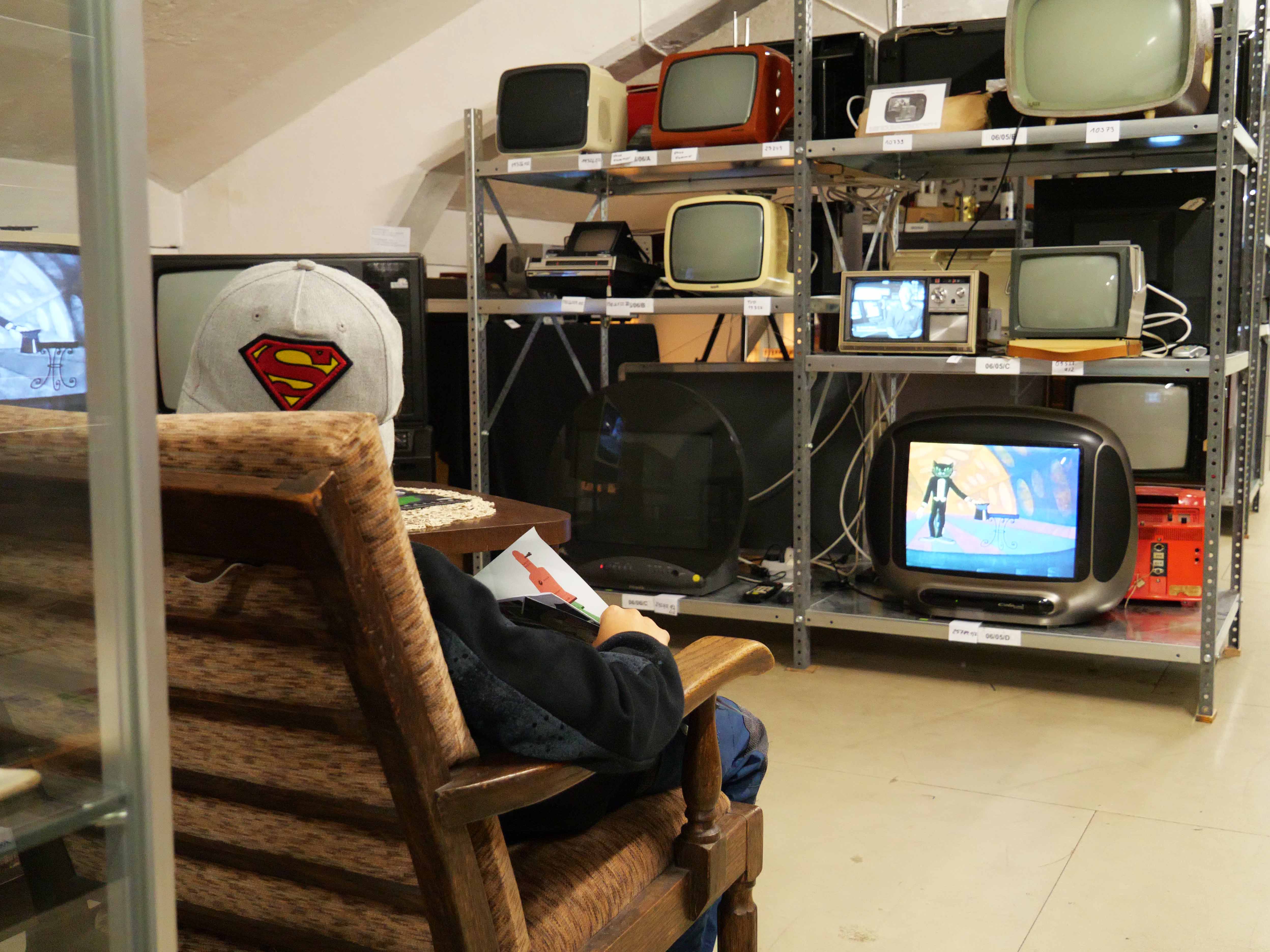 KI generiert: Ein Kind sitzt auf einem Stuhl und schaut auf ein Regal mit alten Fernsehern, von denen einige eingeschaltet sind. Das Bild zeigt eine nostalgische Sammlung von Retro-Fernsehern in einem Raum.