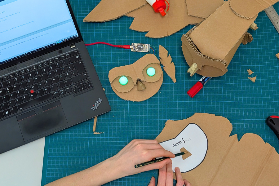 AI generated: A craft project is underway in which someone is creating an owl's head with glowing eyes out of cardboard and using a laptop for support. Various tools and materials are scattered around the table.