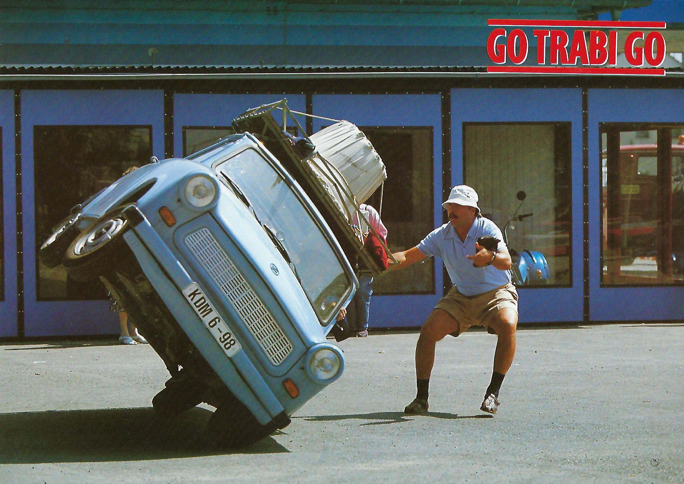 KI generiert: Ein Mann stützt ein blaues Auto, das auf zwei Rädern kippt, während die Aufschrift "Go Trabi Go" im Hintergrund zu sehen ist. Der Hauptinhalt des Bildes ist die humorvolle und dynamische Darstellung eines einradigen Fahrmanövers.