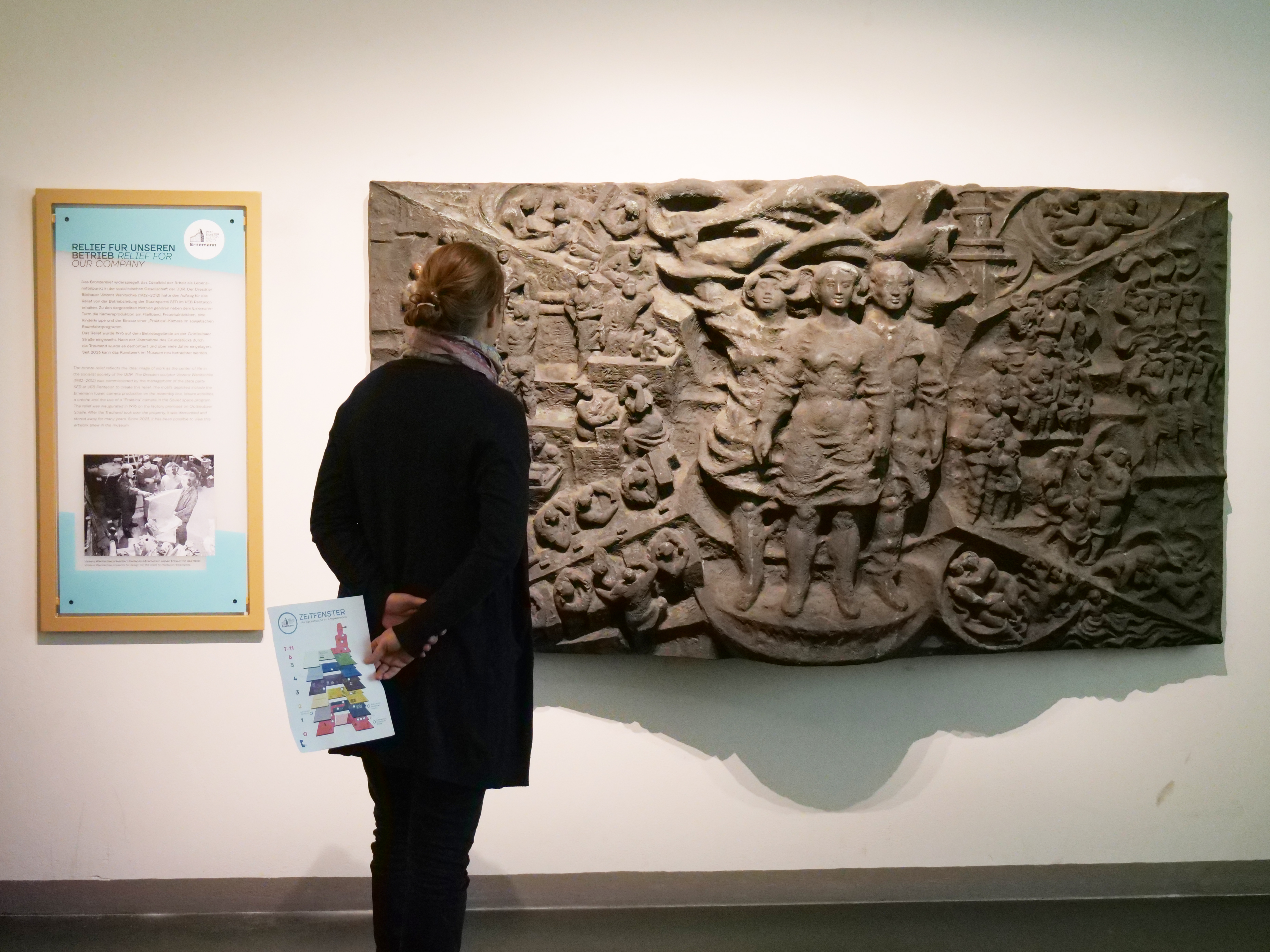 KI generiert: Eine Person betrachtet ein kunstvolles Relief mit verschiedenen Figuren und Motiven an einer Wand. Daneben befindet sich eine Informationstafel über das Kunstwerk.