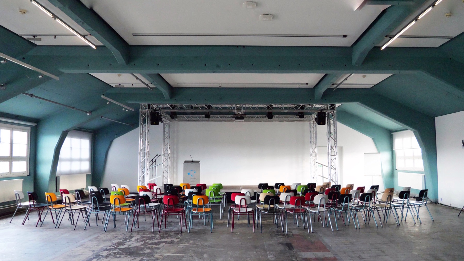 KI generiert: Das Bild zeigt einen Konferenz- oder Veranstaltungsraum mit einer Bühne im Hintergrund und zahlreichen leeren, bunten Stühlen davor. Der Raum ist gut beleuchtet durch mehrere Fenster an den Seiten.