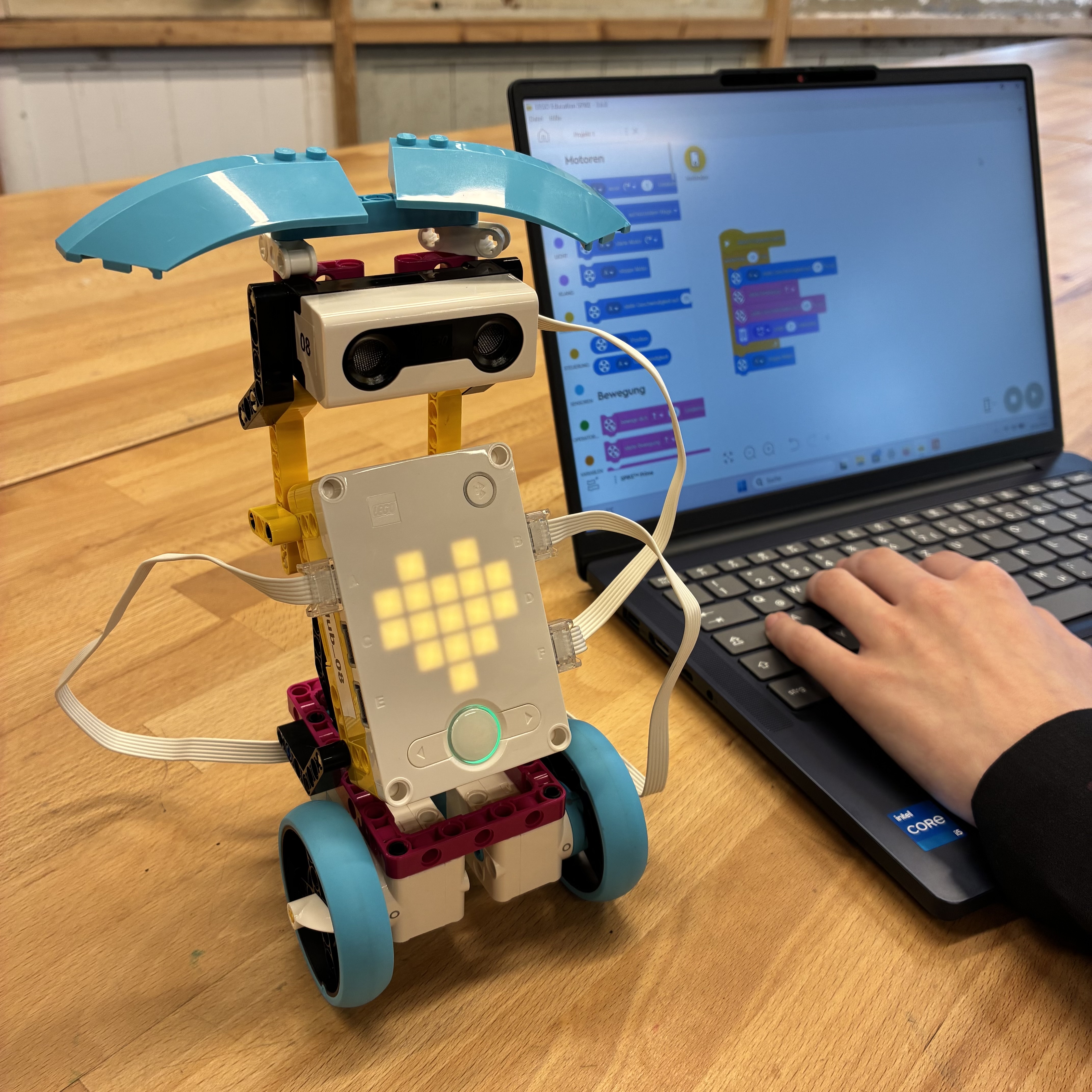 KI generiert: Das Bild zeigt einen spielzeughaften Roboter mit leuchtendem Bildschirm, der ein Herz anzeigt, neben einem Laptop, auf dem ein Programmierinterface zu sehen ist. Eine Person benutzt den Laptop, vermutlich um den Roboter zu programmieren.