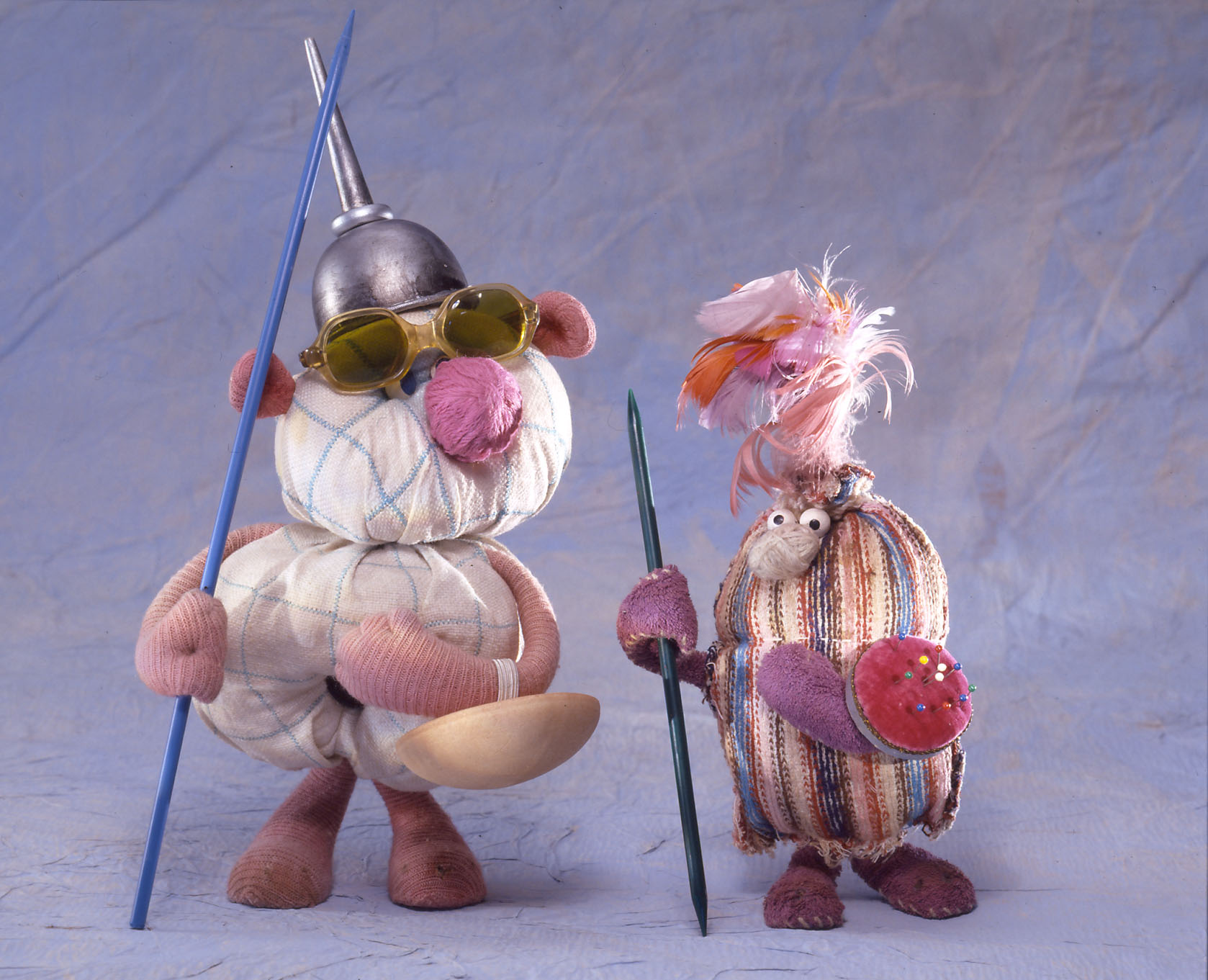 KI generiert: Das Bild zeigt zwei fantasievolle, bunte Stoffpuppen mit skurrilen Accessoires wie Hüten und Speeren auf einem blauen Hintergrund. Die Puppen sind unterschiedlicher Form und Größe und tragen kreative Dekorationen.