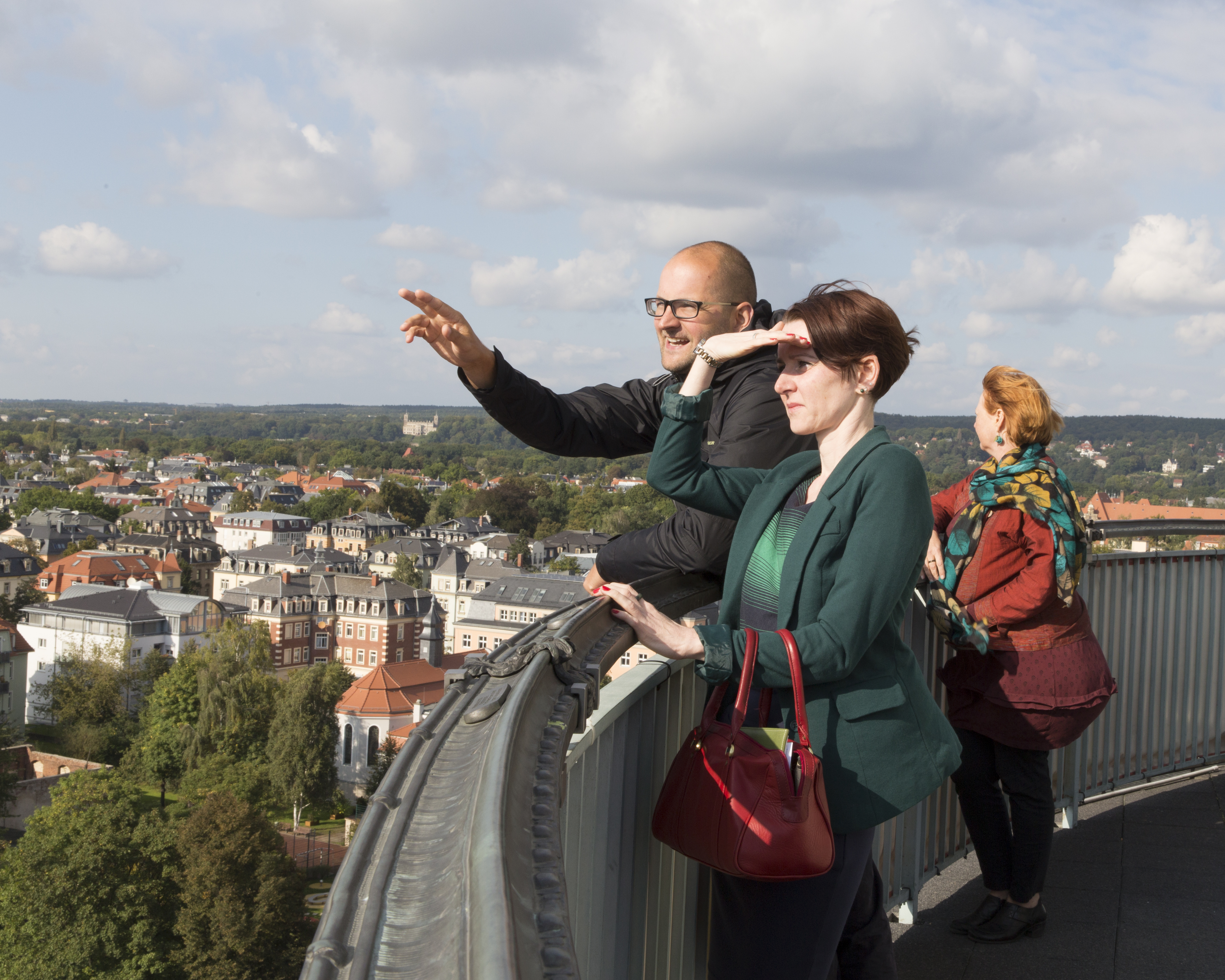 KI generiert: Das Bild zeigt drei Personen, die von einer Aussichtsplattform aus auf eine Stadtlandschaft blicken und die Aussicht genießen. Eine Person im Vordergrund zeigt nach vorne, möglicherweise um anderen etwas Bestimmtes in der Ferne zu zeigen.