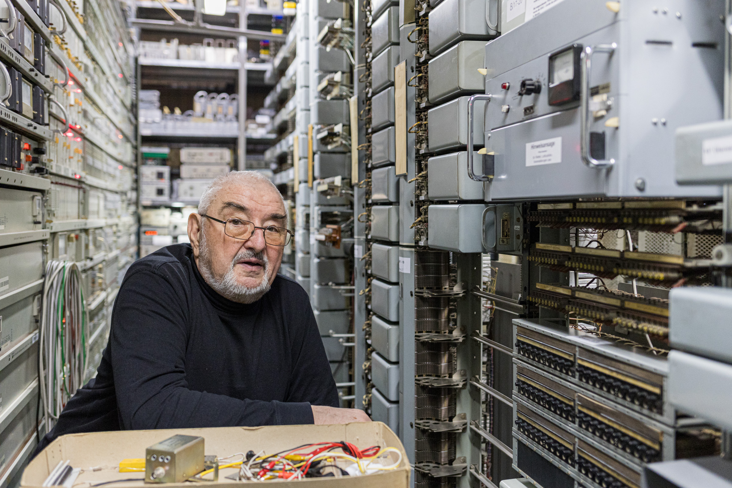 KI generiert: Das Bild zeigt einen älteren Mann, der zwischen zwei Reihen von elektronischen Geräten in einer Art Technik- oder Serverraum steht oder arbeitet. Er blickt in die Kamera und scheint von zahlreichen Kabeln und Hardware umgeben zu sein.