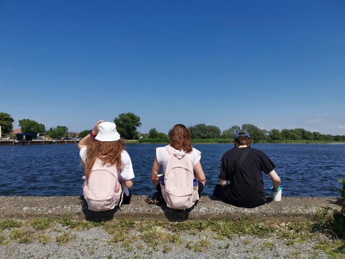 KI generiert: Das Bild zeigt drei Personen, die am Ufer eines Gewässers sitzen, mit der Aussicht auf ruhiges, blaues Wasser und einen klaren Himmel. Sie tragen Rucksäcke und genießen offensichtlich die Umgebung in ruhiger Atmosphäre.