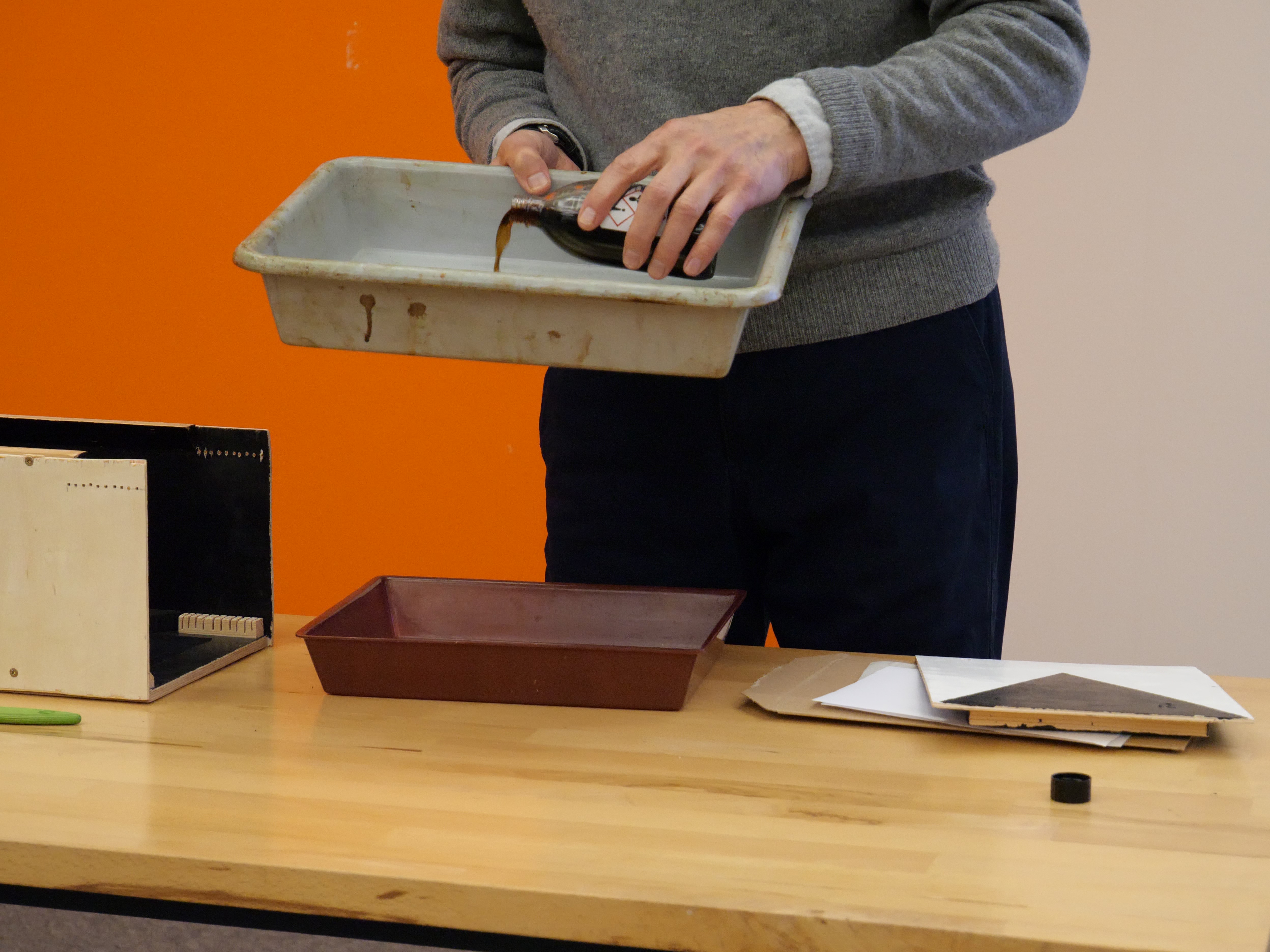 AI generated: A person is apparently preparing a photographic development by pouring a liquid from a bottle into a developer tray. Other utensils for the development process are spread out on the table.