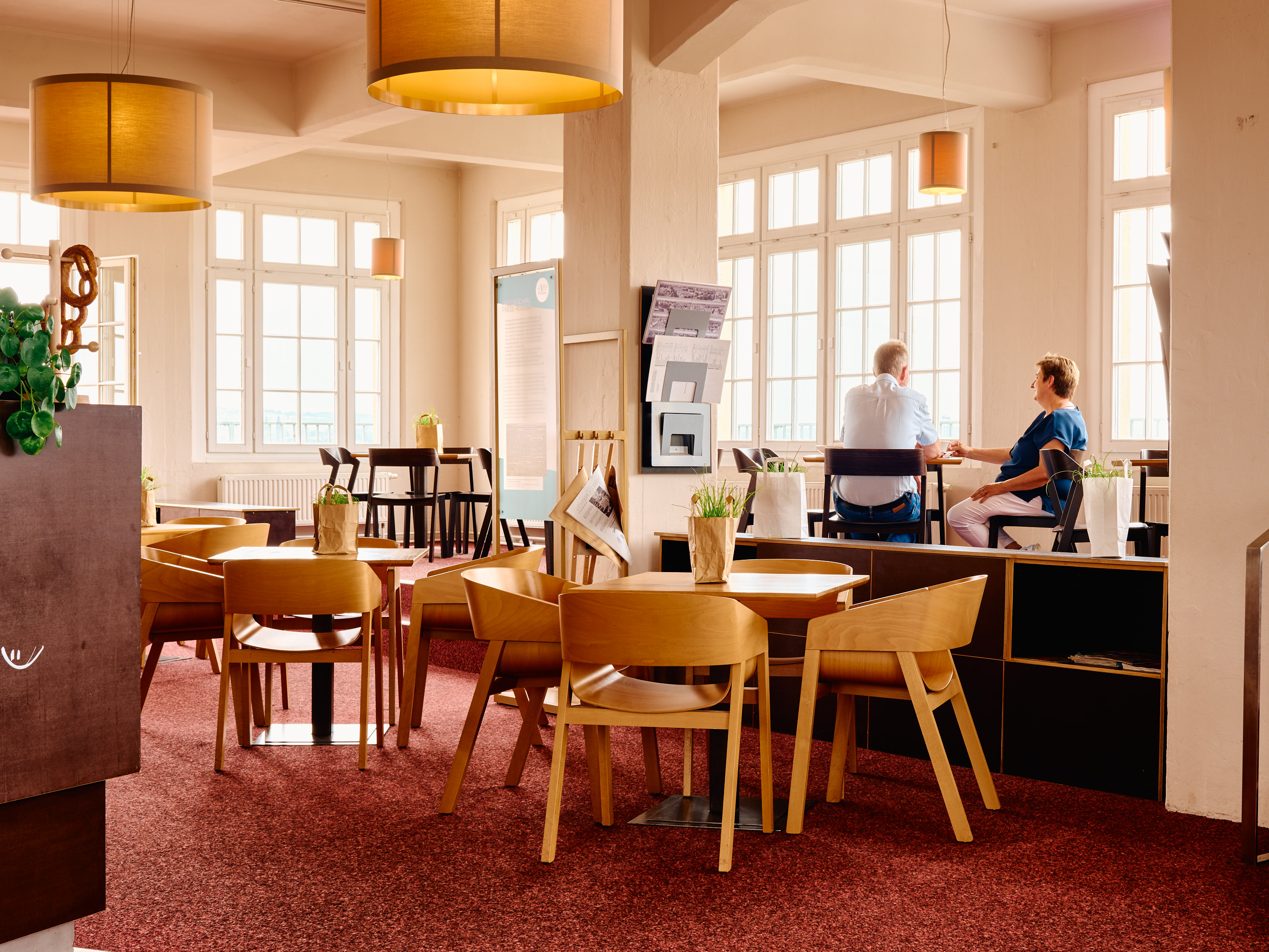 KI generiert: Das Bild zeigt ein gemütliches Café mit Holzmöbeln und großen Fenstern, durch die viel Licht einfällt. Im Hintergrund sitzen zwei Personen an einem Tisch im Gespräch.