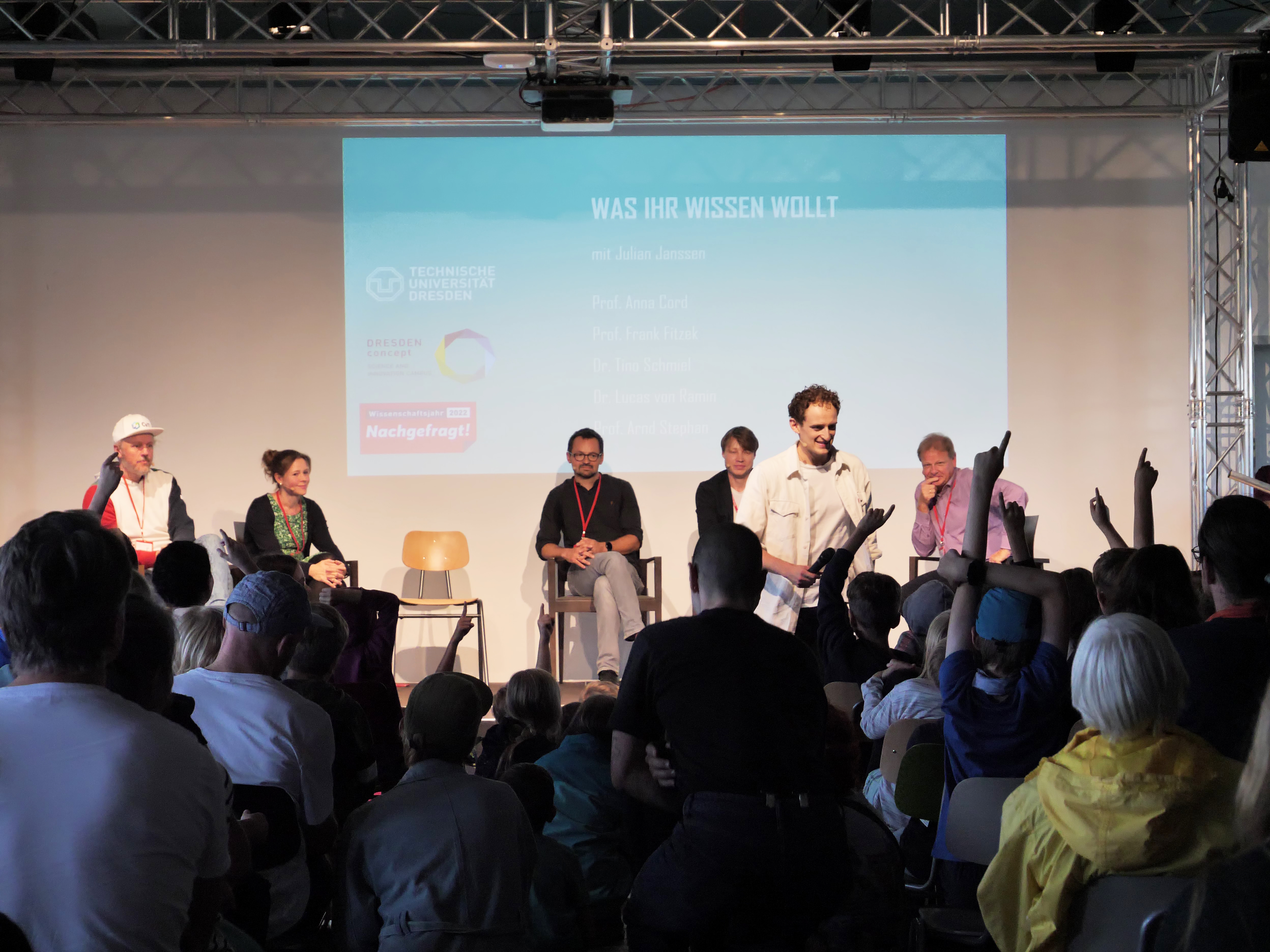 KI generiert: Das Bild zeigt eine Podiumsdiskussion mit mehreren Teilnehmern auf einer Bühne vor einem Publikum. Im Hintergrund wird eine Projektion angezeigt, die das Thema der Veranstaltung und die Namen der Podiumsteilnehmer zeigt.