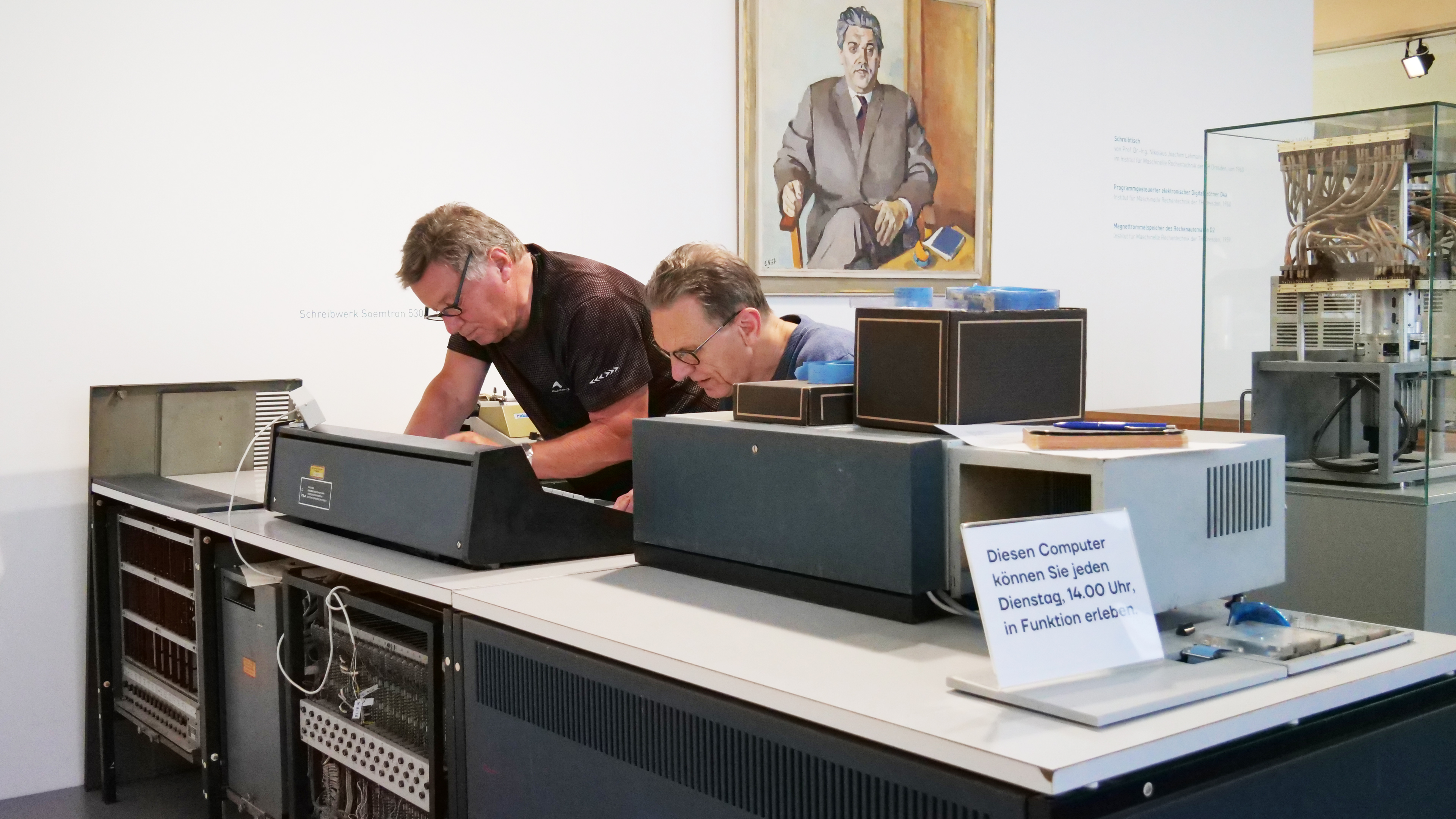 KI generiert: Zwei Männer arbeiten an einem großen alten Computer in einem Museum oder einer Ausstellung. Ein Schild informiert darüber, dass der Computer jeden Dienstag um 14:00 Uhr in Betrieb gezeigt wird.