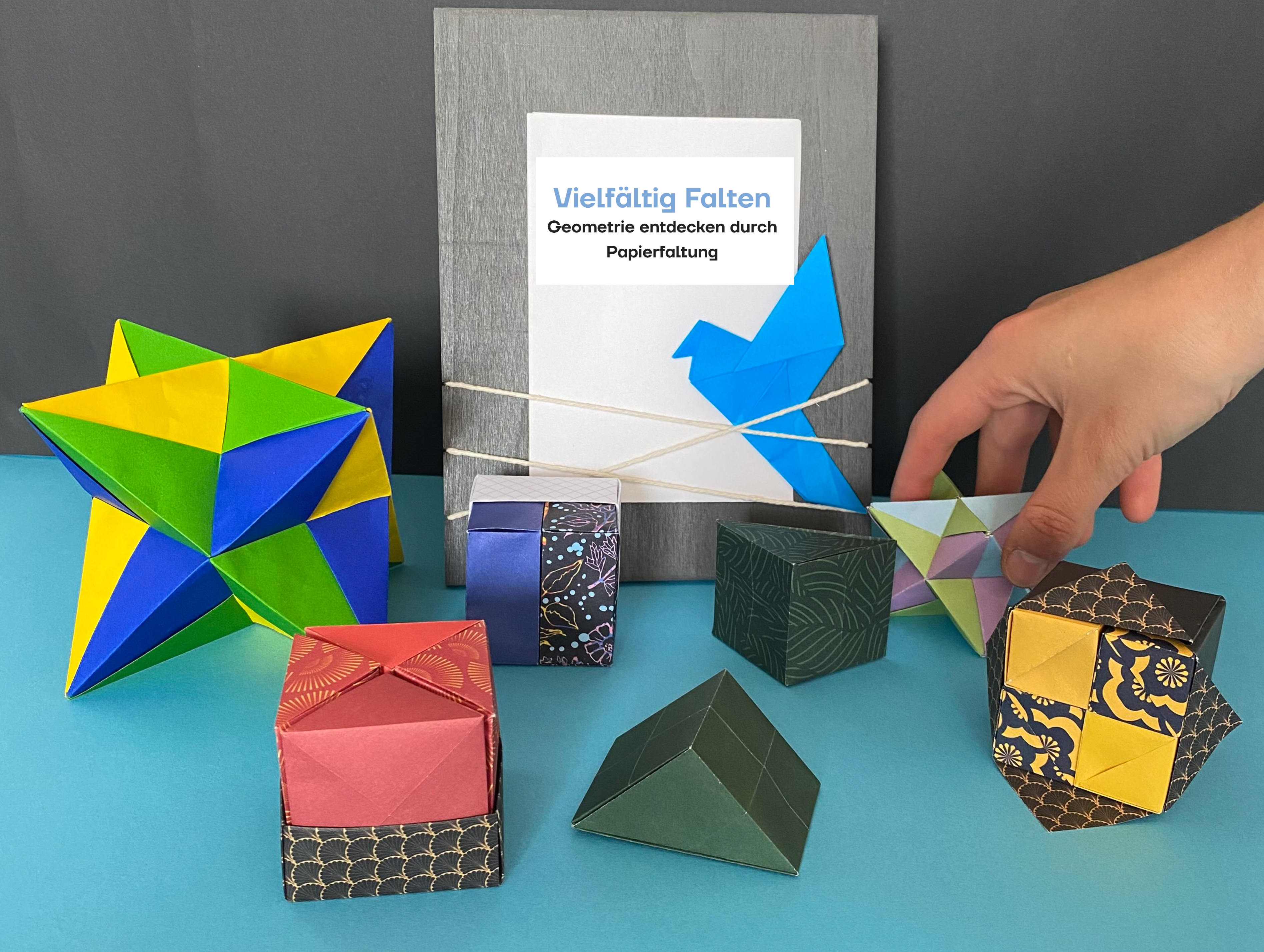 KI generiert: Das Bild zeigt eine Sammlung von bunten, geometrischen Origami-Figuren und eine Informationsbroschüre über das Entdecken von Geometrie durch Papierfalten. Eine Hand greift nach einem der Objekte, umgeben von verschiedenen gefalteten Modellen.