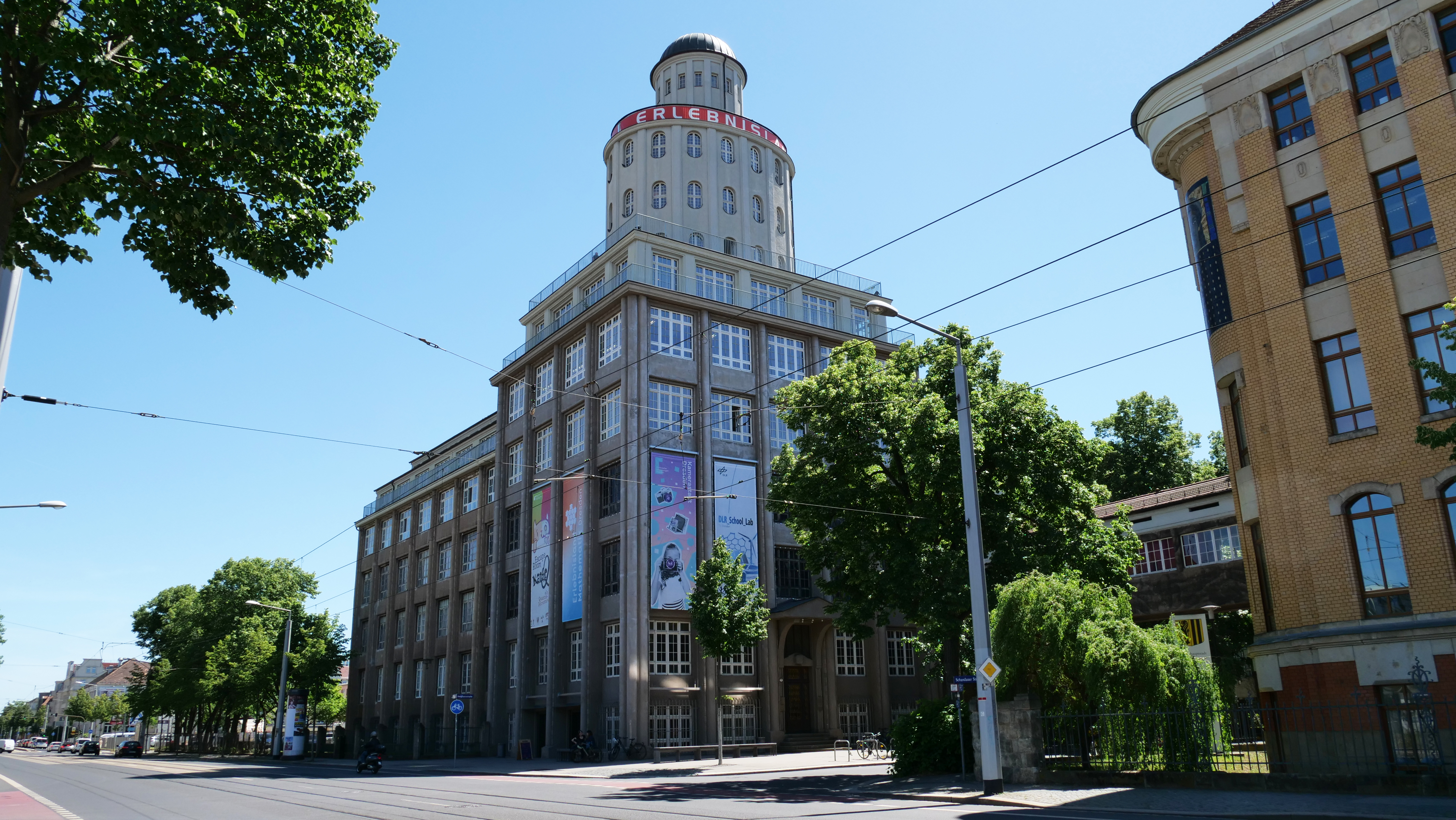 KI generiert: Das Bild zeigt ein großes, historisches Gebäude mit einer markanten runden Turmstruktur und der Aufschrift "Erlebnis". Es ist von Bäumen umgeben und steht entlang einer Straßenbahnlinie unter einem klaren blauen Himmel.